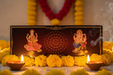 Decorative setup with Lord Ganesha and Goddess Lakshmi on a black background, surrounded by yellow flowers and lit candles.