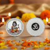 Two silver coins with one featuring a deity and the other an Om symbol, on a decorative background with flowers and candles.